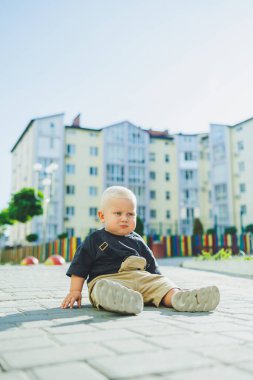 Çocuk parkında açık havada yaz elbisesi giymiş bir çocuk. Bir yaşında bir çocuk, yüksek binaların yakınındaki modern bir avluda yürüyor. Çok katlı bir binada çocuk parkı.