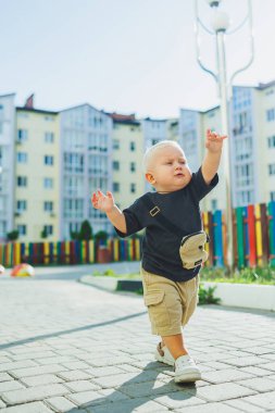 Çocuk parkında açık havada yaz elbisesi giymiş bir çocuk. Bir yaşında bir çocuk, yüksek binaların yakınındaki modern bir avluda yürüyor. Çok katlı bir binada çocuk parkı.