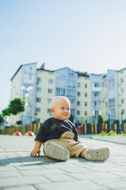 Çocuk parkında açık havada yaz elbisesi giymiş bir çocuk. Bir yaşında bir çocuk, yüksek binaların yakınındaki modern bir avluda yürüyor. Çok katlı bir binada çocuk parkı.
