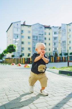 Çocuk parkında açık havada yaz elbisesi giymiş bir çocuk. Bir yaşında bir çocuk, yüksek binaların yakınındaki modern bir avluda yürüyor. Çok katlı bir binada çocuk parkı.