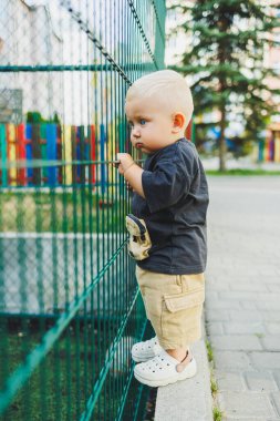 Çocuk parkında açık havada yaz elbisesi giymiş bir çocuk. Bir yaşında bir çocuk, yüksek binaların yakınındaki modern bir avluda yürüyor. Çok katlı bir binada çocuk parkı.