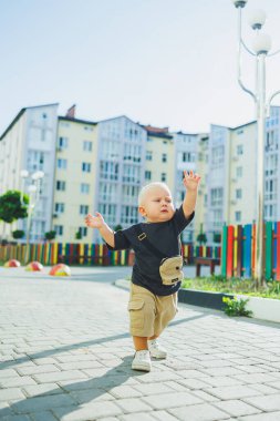 Çocuk parkında açık havada yaz elbisesi giymiş bir çocuk. Bir yaşında bir çocuk, yüksek binaların yakınındaki modern bir avluda yürüyor. Çok katlı bir binada çocuk parkı.
