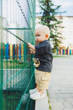 Çocuk parkında açık havada yaz elbisesi giymiş bir çocuk. Bir yaşında bir çocuk, yüksek binaların yakınındaki modern bir avluda yürüyor. Çok katlı bir binada çocuk parkı.