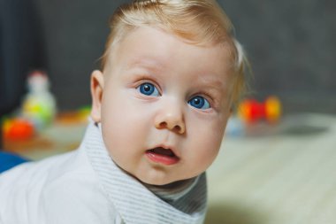 Mavi gözlü bir bebek yerde yatıyor ve başını tutuyor. Bebeğin gelişimi. Beyaz elbiseli bir çocuk.