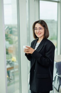 Gözlüklü güzel bir kadın şeffaf bir ofiste pencerenin yanında duruyor. Ofisteki pencereden bakan bir kadın dinleniyor.
