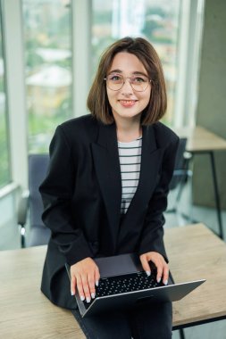 Ofiste dizüstü bilgisayarla çalışan kadın yönetici gülümsüyor. Bir çalışan büyük pencereli bir ofiste çalışır..