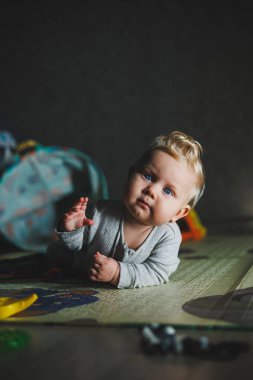 Evde bebek oyuncaklarıyla oynayan tatlı sarışın çocuk oturma odasında halının üzerinde oturuyor. Gülümseyen küçük sevimli çocuk renkli oyuncaklar kullanıyor, ip çiğniyor.