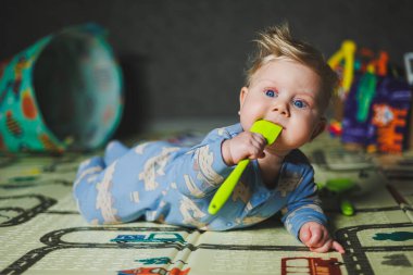 Evde bebek oyuncaklarıyla oynayan tatlı sarışın çocuk oturma odasında halının üzerinde oturuyor. Gülümseyen küçük sevimli çocuk renkli oyuncaklar kullanıyor, ip çiğniyor.