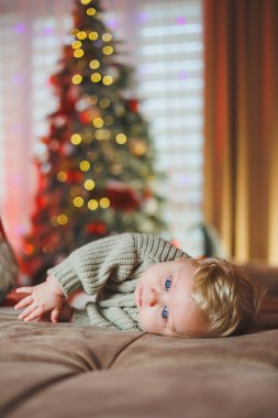 Noel ağacı oyuncaklarıyla arka planda sevimli bir erkek bebek, geleneksel Noel kostümü. Yeni yıl tatili. Bir bebek için ilk yeni yıl