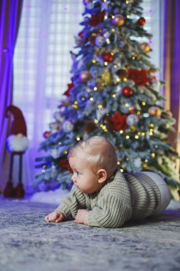 Noel ağacının arka planında bej örülmüş süveter giyen bir bebek. Bebeğin ilk Noel 'i. Noel süslemelerinin arka planında küçük bir çocuk.