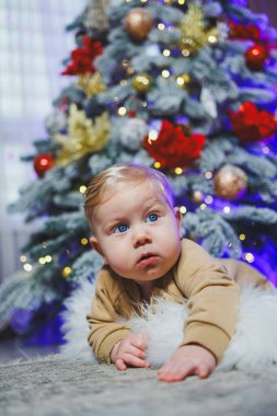 Mavi gözlü bir bebek bir Noel ağacının arka planında yerde yatıyor. Noel atmosferinde evde bir çocuk. Bebeğin ilk yeni yılı