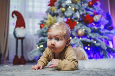 Mavi gözlü bir bebek bir Noel ağacının arka planında yerde yatıyor. Noel atmosferinde evde bir çocuk. Bebeğin ilk yeni yılı