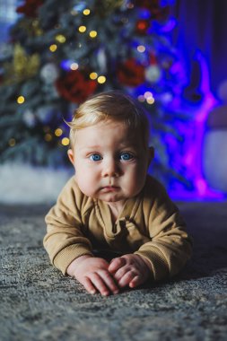 Mavi gözlü bir bebek bir Noel ağacının arka planında yerde yatıyor. Noel atmosferinde evde bir çocuk. Bebeğin ilk yeni yılı