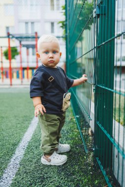 Futbol sahasında tişört ve pantolon giymiş 1 yaşında küçük bir çocuk koşuyor. Bir çocuk dışarıda oynuyor. Şık çocuk kıyafetleri içindeki sarışın bebek..