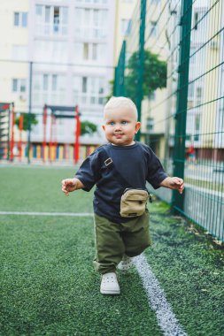 Futbol sahasında tişört ve pantolon giymiş 1 yaşında küçük bir çocuk koşuyor. Bir çocuk dışarıda oynuyor. Şık çocuk kıyafetleri içindeki sarışın bebek..