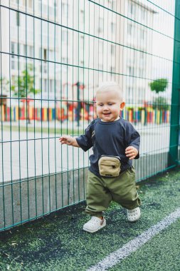 A small 1-year-old boy in a T-shirt and pants is running on the football field. A child is playing outdoors. Blond baby in stylish children's clothes. clipart