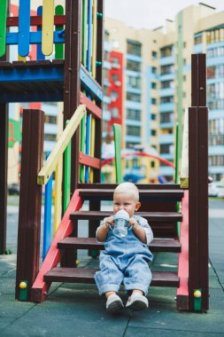 Kot pantolon giyen bir yaşındaki bebek sokakta bir şişeden su içiyor. Büyük boy çocuk tulumu. Çocuk parkında şık küçük bir çocuk.