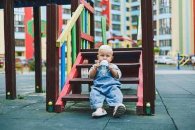 Kot pantolon giyen bir yaşındaki bebek sokakta bir şişeden su içiyor. Büyük boy çocuk tulumu. Çocuk parkında şık küçük bir çocuk.