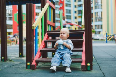 Kot pantolon giyen bir yaşındaki bebek sokakta bir şişeden su içiyor. Büyük boy çocuk tulumu. Çocuk parkında şık küçük bir çocuk.