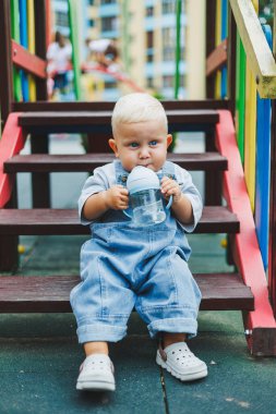 Kot pantolon giyen bir yaşındaki bebek sokakta bir şişeden su içiyor. Büyük boy çocuk tulumu. Çocuk parkında şık küçük bir çocuk.