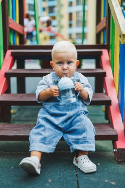 Kot pantolon giyen bir yaşındaki bebek sokakta bir şişeden su içiyor. Büyük boy çocuk tulumu. Çocuk parkında şık küçük bir çocuk.