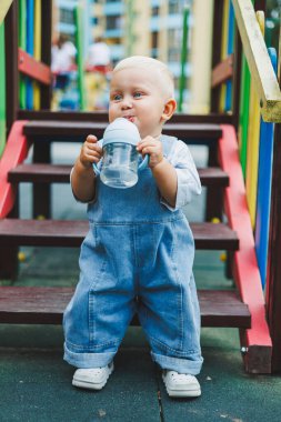 Kot pantolon giyen bir yaşındaki bebek sokakta bir şişeden su içiyor. Büyük boy çocuk tulumu. Çocuk parkında şık küçük bir çocuk.