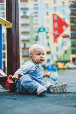 Kot pantolon giyen bir yaşındaki bebek sokakta bir şişeden su içiyor. Büyük boy çocuk tulumu. Çocuk parkında şık küçük bir çocuk.