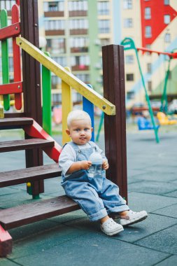 Kot pantolon giyen bir yaşındaki bebek sokakta bir şişeden su içiyor. Büyük boy çocuk tulumu. Çocuk parkında şık küçük bir çocuk.