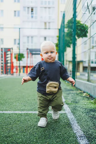 Futbol sahasında tişört ve pantolon giymiş 1 yaşında küçük bir çocuk koşuyor. Bir çocuk dışarıda oynuyor. Şık çocuk kıyafetleri içindeki sarışın bebek..