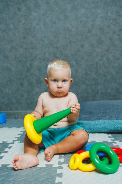 Şortlu küçük bir çocuk evde halıya oturup eğitici oyuncaklarla oynuyor. Bir çocuk plastik piramit inşa eder..