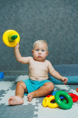 Şortlu küçük bir çocuk evde halıya oturup eğitici oyuncaklarla oynuyor. Bir çocuk plastik piramit inşa eder..