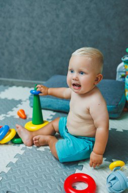Şortlu küçük bir çocuk evde halıya oturup eğitici oyuncaklarla oynuyor. Bir çocuk plastik piramit inşa eder..