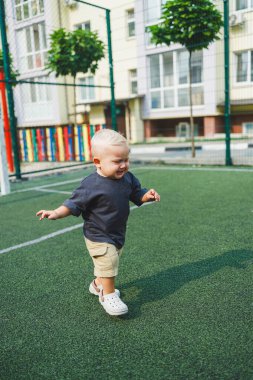 Tişört ve şort giyen 1 yaşında sarışın bir çocuk evin bahçesindeki spor sahasında koşuyor. Bir çocuk dışarıda oynuyor. Erkekler için şık kıyafetler. Yaz çocuk ayakkabıları..