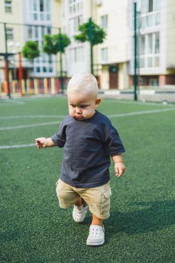 Tişört ve şort giyen 1 yaşında sarışın bir çocuk evin bahçesindeki spor sahasında koşuyor. Bir çocuk dışarıda oynuyor. Erkekler için şık kıyafetler. Yaz çocuk ayakkabıları..
