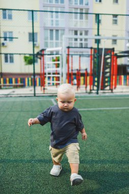 Tişört ve şort giyen 1 yaşında sarışın bir çocuk evin bahçesindeki spor sahasında koşuyor. Bir çocuk dışarıda oynuyor. Erkekler için şık kıyafetler. Yaz çocuk ayakkabıları..