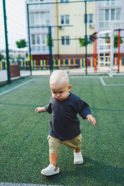 Tişört ve şort giyen 1 yaşında sarışın bir çocuk evin bahçesindeki spor sahasında koşuyor. Bir çocuk dışarıda oynuyor. Erkekler için şık kıyafetler. Yaz çocuk ayakkabıları..