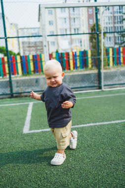 Parktaki stadyumda koşan neşeli 1 yaşında bir çocuk. Yeşil çim tarlası. Çocuğun aktif yaşam tarzı bu. Bir bebek için sağlıklı bir yaşam tarzı. Bir erkek için şık kıyafetler..