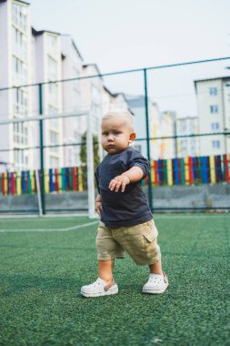 Parktaki stadyumda koşan neşeli 1 yaşında bir çocuk. Yeşil çim tarlası. Çocuğun aktif yaşam tarzı bu. Bir bebek için sağlıklı bir yaşam tarzı. Bir erkek için şık kıyafetler..