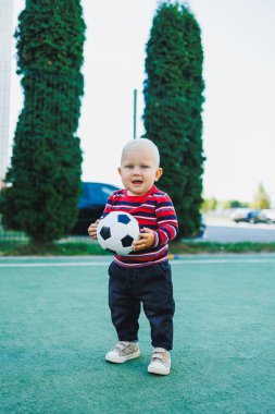 Bir yaşında bir çocuk sokakta bir futbol topuyla oynuyor. Çocuk temiz havada oyun oynuyor. Şık küçük sarışın çocuk..