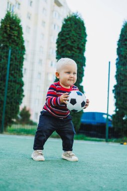 Bir yaşında bir çocuk sokakta bir futbol topuyla oynuyor. Çocuk temiz havada oyun oynuyor. Şık küçük sarışın çocuk..