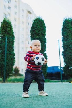 Bir yaşında bir çocuk sokakta bir futbol topuyla oynuyor. Çocuk temiz havada oyun oynuyor. Şık küçük sarışın çocuk..