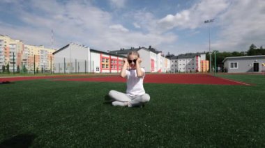 Güneş gözlüklü küçük bir kız parkta koşuyor ve gülümsüyor. Beyaz tişörtlü şık bir kız. Saçları örgülü bir kız.
