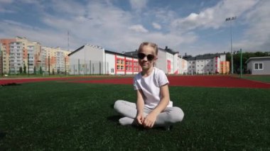 Güneş gözlüklü küçük bir kız parkta koşuyor ve gülümsüyor. Beyaz tişörtlü şık bir kız. Saçları örgülü bir kız.