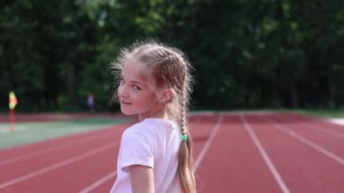 Stadyumda koşan küçük bir kız, atılan havada koşu bandında koşuyor. Beyaz tişörtlü şık bir kız. Saçları örgülü bir kız.