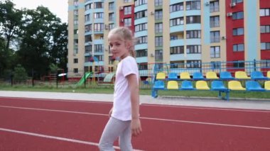 Stadyumda koşan küçük bir kız, atılan havada koşu bandında koşuyor. Beyaz tişörtlü şık bir kız. Saçları örgülü bir kız.