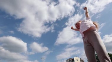 Küçük bir kız gülüyor ve mavi gökyüzünün arka planında dans ediyor. Beyaz tişörtlü şık bir kız. Saçları örgülü bir kız.