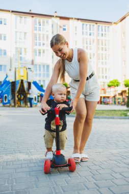 Bir anne küçük oğluna scooter sürmeyi öğretir. Sokakta anne ve çocuk oyunları. Aile yürüyüşü kavramı.