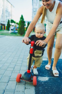 Bir anne küçük oğluna scooter sürmeyi öğretir. Sokakta anne ve çocuk oyunları. Aile yürüyüşü kavramı.