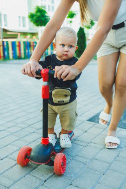Bir anne küçük oğluna scooter sürmeyi öğretir. Sokakta anne ve çocuk oyunları. Aile yürüyüşü kavramı.