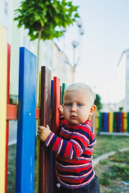 Küçük sarışın bir çocuk tahta bir çitin yanında yürür. Çocuk temiz havada oyun oynuyor. Çocuk gelişimi. Kot ve spor ayakkabılı bir çocuk..